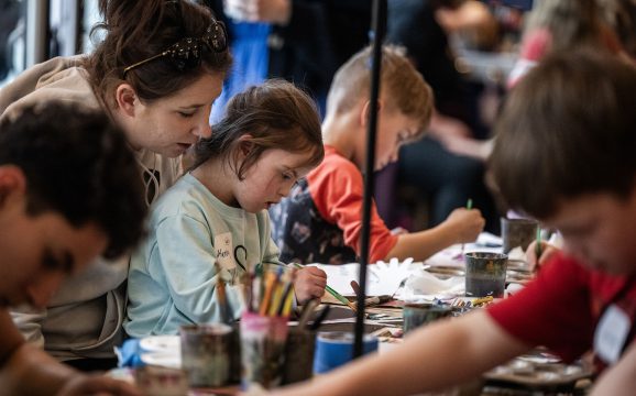 Women helps girl work on art project at table with other kids